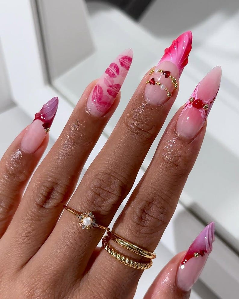 May include: A hand displaying long, almond-shaped acrylic nails. The nails are decorated with designs such as pink lips, red hearts, and gold detailing. Two gold rings are visible on the ring finger.