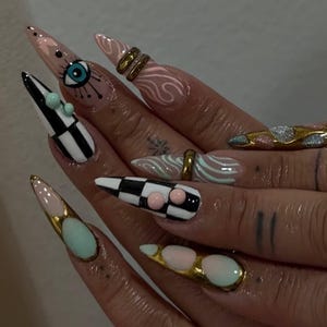Peut inclure: Gros plan sur des ongles longs et pointus avec un nail art &eacute;labor&eacute;. Les motifs comprennent un motif de damier, un &oelig;il, des volutes et des accents dor&eacute;s. Les ongles sont noirs, blancs, roses, dor&eacute;s et bleus. Des bagues et de petits ornements ornent les ongles.