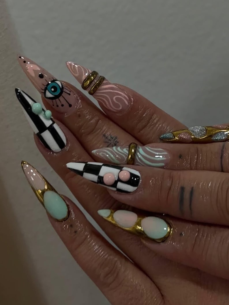 Peut inclure: Gros plan sur des ongles longs et pointus avec un nail art &eacute;labor&eacute;. Les motifs comprennent un motif de damier, un &oelig;il, des volutes et des accents dor&eacute;s. Les ongles sont noirs, blancs, roses, dor&eacute;s et bleus. Des bagues et de petits ornements ornent les ongles.