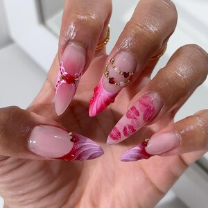 May include: Close-up of long, pointed acrylic nails with various designs. One nail features a French tip with a red and purple swirl, while others showcase pink gradients, heart and bead accents, and intricate patterns. The nails are a mix of pink, red, and white.