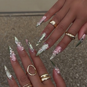 May include: Stiletto-shaped artificial nails with a floral design. The nails feature a gradient of pink and green, adorned with white flowers and silver accents. Gold rings of various designs are also visible.