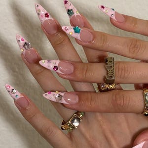 May include: Pointed stiletto nails featuring a light pink French tip design adorned with small colorful star-shaped charms and tiny rhinestones. The fingers are decorated with gold-toned metal rings, including one with engraved text.