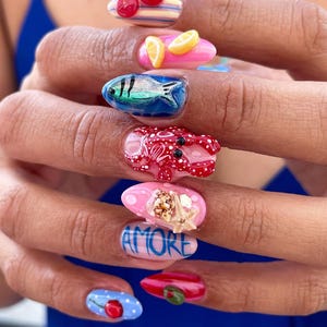 May include: Manicured nails featuring 3D art including red cherry halves, yellow lemon slices, a metallic blue fish, a red lobster, a seashell with a starfish, the word AMORE in blue, a blue nail with a red cherry, and a red nail with a green olive.
