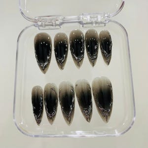 May include: A set of ten long, pointed false nails in a clear plastic case. Each nail features a gradient design, transitioning from a dark, smoky black at the tip to a lighter, smoky gray at the base. Silver, swirling accents add detail.