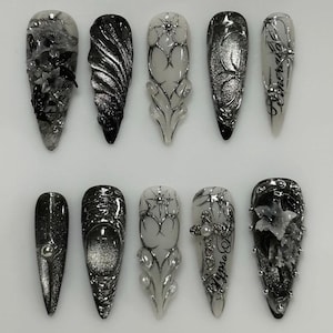May include: A set of ten long, pointed artificial nails. The nails feature intricate designs in black, silver, and white, including floral patterns, geometric shapes, and text. Some nails have a glossy finish, while others have a matte appearance.
