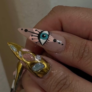 Peut inclure: Gros plan sur deux longs ongles pointus avec du nail art. L'un des ongles pr&eacute;sente un motif m&eacute;tallique dor&eacute; avec des accents de paillettes. L'autre ongle a une base nude avec un motif d'&oelig;il en noir, rose et bleu.