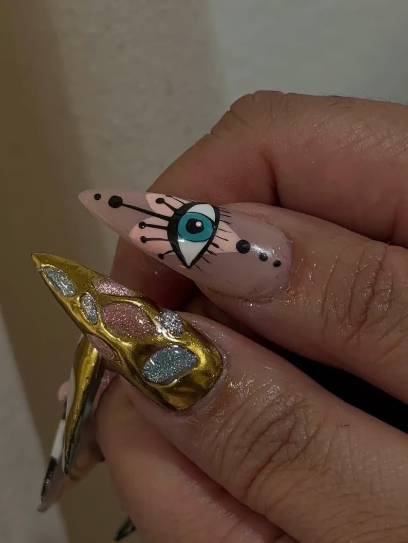 Peut inclure: Gros plan sur deux longs ongles pointus avec du nail art. L'un des ongles pr&eacute;sente un motif m&eacute;tallique dor&eacute; avec des accents de paillettes. L'autre ongle a une base nude avec un motif d'&oelig;il en noir, rose et bleu.