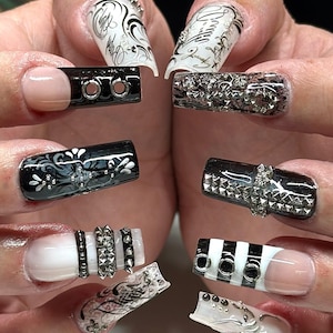 May include: Close-up of manicured nails with various designs. Some nails are black with silver accents, others are white with black stripes or silver studs. Several nails feature intricate patterns and embellishments.