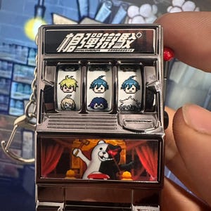 Peut inclure: Un porte-clés en forme de machine à sous argentée avec du texte japonais. Les rouleaux affichent des images de personnages pixélisés. La section inférieure montre un personnage d'ours noir et blanc sur une scène avec des rideaux rouges.