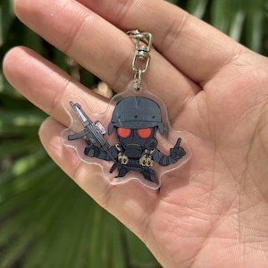May include: A clear acrylic keychain featuring a cartoon soldier in a gas mask and helmet, holding a rifle. The soldier is dressed in black with red eyes. The keychain has a silver metal clasp.