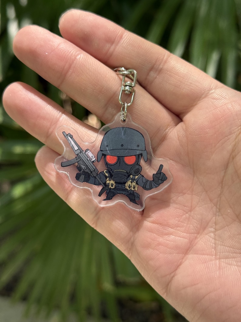 May include: A clear acrylic keychain featuring a cartoon soldier in a gas mask and helmet, holding a rifle. The soldier is dressed in black with red eyes. The keychain has a silver metal clasp.