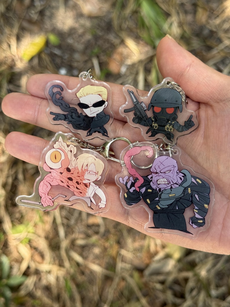 May include: Four acrylic keychains featuring cartoon characters. One keychain depicts a character with sunglasses and a black outfit. Another shows a character in a gas mask holding a gun. The other two keychains show characters with mutated features.