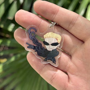 May include: A clear acrylic keychain featuring a cartoon character with blonde hair, sunglasses, and a black outfit. The character is holding a dark, tentacle-like object. The keychain has a silver chain.