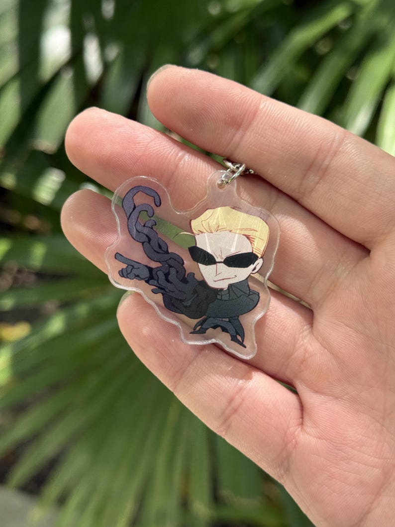 May include: A clear acrylic keychain featuring a cartoon character with blonde hair, sunglasses, and a black outfit. The character is holding a dark, tentacle-like object. The keychain has a silver chain.