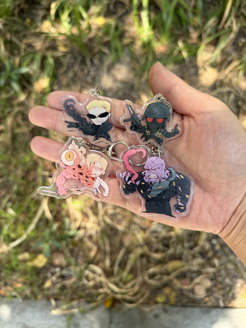 May include: Four acrylic keychains featuring cartoon characters. Each keychain has a different character design, including a character with sunglasses, a soldier with a helmet, a character with a mutated body, and a purple-skinned character.