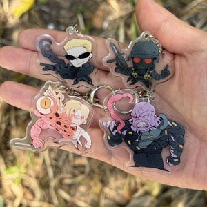 May include: Four clear acrylic keychains featuring cartoon characters. One keychain depicts a character with sunglasses and black clothing. Another shows a character in a helmet holding a gun. The other two keychains show other characters with unique designs.