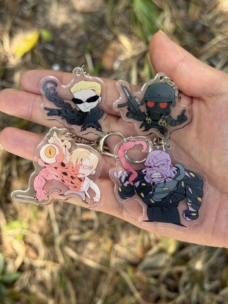 May include: Four clear acrylic keychains featuring cartoon characters. One keychain depicts a character with sunglasses and black clothing. Another shows a character in a helmet holding a gun. The other two keychains show other characters with unique designs.