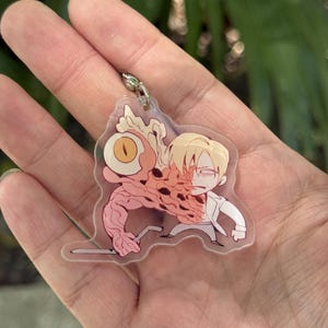 May include: A clear acrylic keychain featuring a cartoon illustration of a character with a large eye and a mutated arm. The character is wearing a white coat and has blonde hair. The keychain has a silver-colored metal clasp.
