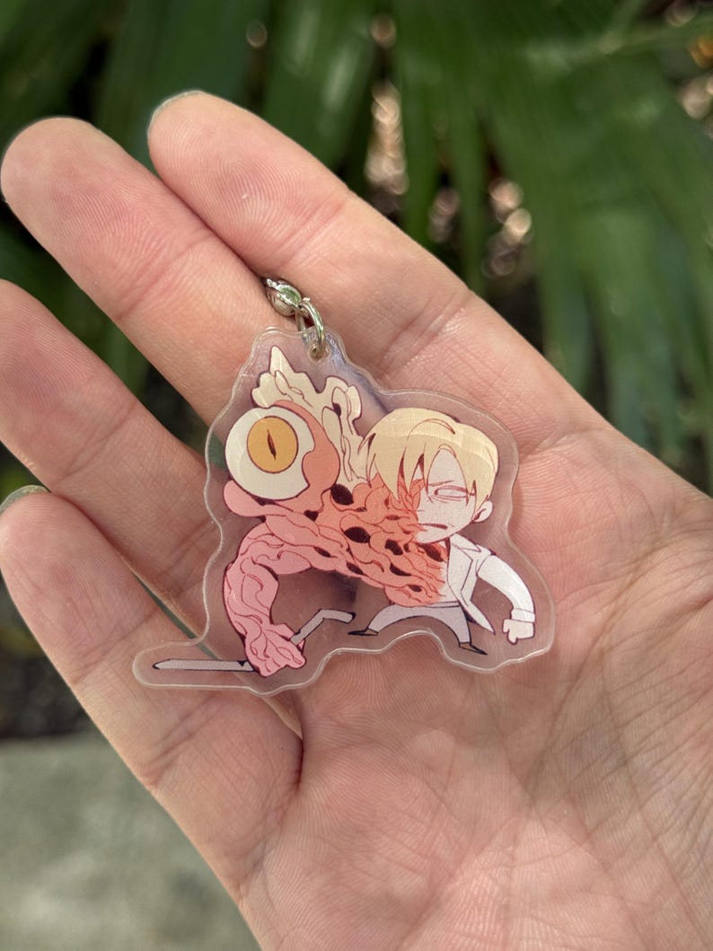 May include: A clear acrylic keychain featuring a cartoon illustration of a character with a large eye and a mutated arm. The character is wearing a white coat and has blonde hair. The keychain has a silver-colored metal clasp.