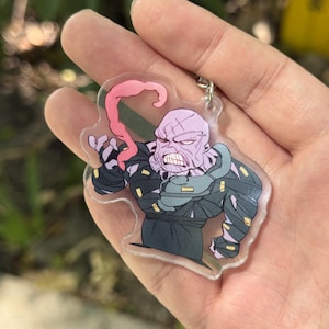 May include: A clear acrylic keychain featuring a cartoon illustration of a muscular, purple-skinned character with a protruding pink tongue. The character is wearing a dark outfit with green accents. The keychain has a silver metal ring.