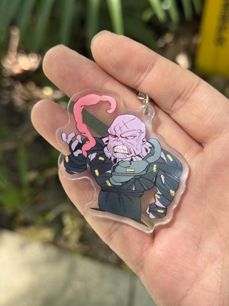 May include: A clear acrylic keychain featuring a cartoon illustration of a muscular, purple-skinned character with a protruding pink tongue. The character is wearing a dark outfit with green accents. The keychain has a silver metal ring.