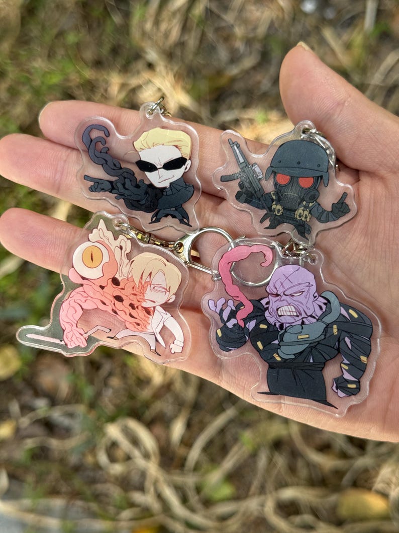 May include: Four acrylic keychains featuring cartoon characters. The keychains depict characters with various designs, including a character with sunglasses and a black outfit, a character with a gas mask and a gun, and two other unique characters.