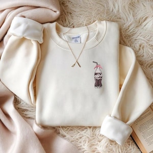May include: Cream-colored sweatshirt featuring an embroidered Dr. Pepper bottle with a pink bow and straw. A gold necklace with a pendant is draped on the shirt. The sleeves are cuffed, and the garment is displayed on a textured surface.