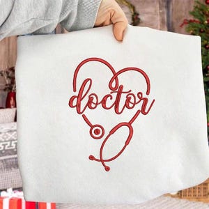 Puede incluir: Sudadera blanca con un diseño rojo en forma de corazón. La palabra "doctor" está escrita en cursiva dentro del corazón, con un estetoscopio incorporado en el diseño. La sudadera es sostenida por una persona.