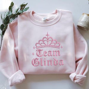 May include: A light pink sweatshirt with the embroidered text "Team Glinda" in a gothic font, topped with a pink crown design. The sweatshirt has a crew neck and long sleeves. A sprig of greenery and a ceramic mug are in the background.