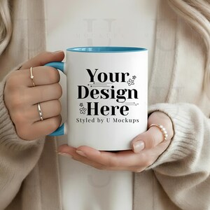 Op de afbeelding: Een witte keramische mok met een blauwe binnenkant en handvat, met de zwarte tekst "Your Design Here". De mok wordt vastgehouden door een persoon die een beige vest en ringen draagt.
