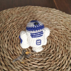 May include: A crocheted R2-D2 robot toy in white and blue with a black eye. The toy is sitting on a woven straw mat.