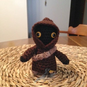 Puede incluir: Una figura de crochet marrón con capucha y ojos amarillos. La figura lleva una bufanda a rayas marrón y beige. La figura está de pie sobre una estera de paja tejida.