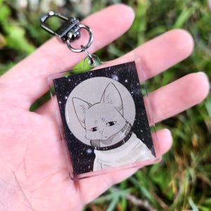 May include: A clear acrylic keychain featuring a gray illustration of a cat in a space suit against a starry background. The cat is centered in front of a large, gray moon. The keychain has a silver clasp.