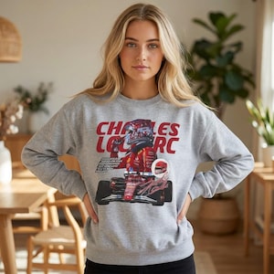 Może przedstawiać: Szara bluza z nadrukiem czerwonego samochodu wyścigowego i napisem "CHARLES LECLERC". Projekt zawiera kierowcę w kasku i podpis. Bluza jest w stylu casual.
