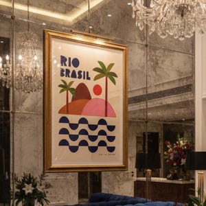 Peut inclure: Un hall d'hôtel élégant avec un canapé bleu capitonné, une œuvre d'art encadrée d'or avec le texte "RIO BRASIL" et un lustre en cristal. L'œuvre représente un paysage coloré avec des palmiers et des vagues. Des compositions florales et des murs en miroir ajoutent à l'ambiance.