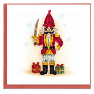 Peut inclure: Une œuvre d'art en papier quilling représentant un soldat casse-noisette avec un chapeau rouge, une veste rouge à accents jaunes et des bottes noires. La figurine tient une épée et se tient entre deux petits cadeaux, un rouge et un vert.