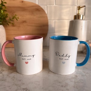 Puede incluir: Dos tazas de cerámica blanca con interior y asa de color. Una taza tiene el interior y el asa de color rosa con el texto Mummy EST. 2016 y un corazón rosa. La otra taza tiene el interior y el asa de color azul con el texto Daddy EST. 2016 y un corazón azul.