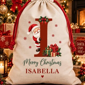 Pode incluir: Um saco de presente bege com cordões vermelhos. A frente apresenta uma letra I vermelha grande, um Papai Noel em desenho animado, uma pilha de presentes embrulhados e o texto Merry Christmas ISABELLA em vermelho e verde com um pequeno coração.