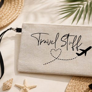 Könnte beinhalten: Eine rechteckige beigefarbene Stofftasche mit schwarzer Handschlaufe und Reißverschluss. Auf der Vorderseite befindet sich der schwarze Schriftzug Travel Stuff, ein gepunktetes Herz und die Silhouette eines Flugzeugs auf einer gestrichelten Linie.