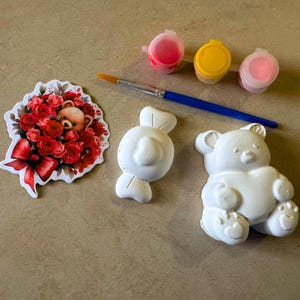 May include: A craft kit featuring a bouquet sticker, a white candy-shaped figurine, and a white teddy bear figurine. The kit includes a paintbrush and three small paint pots in pink and yellow.