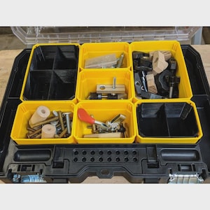Peut inclure: Une mallette de rangement à outils en plastique noir comprenant six bacs amovibles jaunes et noirs. Les bacs contiennent divers composants de quincaillerie, dont des boulons métalliques, des chevilles en bois, une sauterelle à levier rouge.
