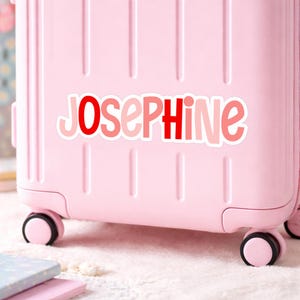 K&ouml;nnte beinhalten: Ein hellrosa Koffer mit einem wei&szlig;en Aufkleber, auf dem der Name JOSEPHINE in einer verspielten, abgerundeten Schrift steht. Die Buchstaben wechseln zwischen leuchtendem Rot und zartem Rosa und sind von einer wei&szlig;en Kontur umgeben.
