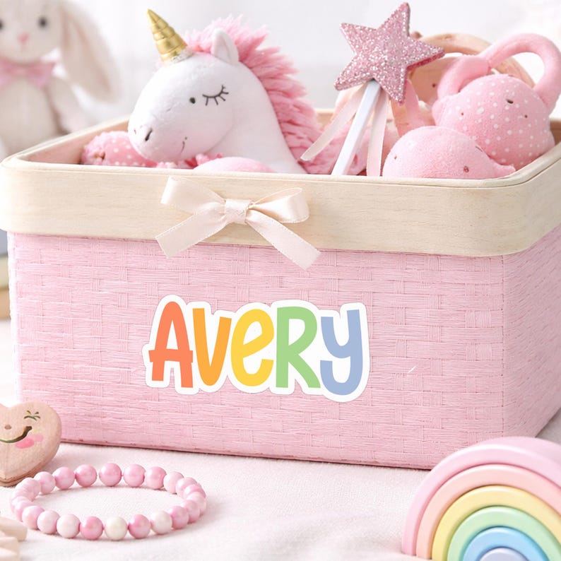 K&ouml;nnte beinhalten: Ein rechteckiger, rosa geflochtener Aufbewahrungskorb mit hellem Holzrand und einer kleinen beigen Schleife. Auf der Vorderseite steht der Name AVERY in bunten, abgerundeten Buchstaben. Darin befinden sich Pl&uuml;schtiere, darunter ein wei&szlig;es Einhorn mit Goldhorn.