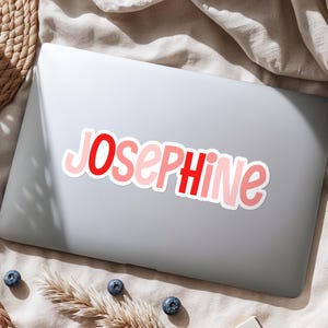 K&ouml;nnte beinhalten: Ein gestanzter Vinyl-Aufkleber mit dem Namen Josephine in einer verspielten, abgerundeten Schriftart. Die Buchstaben wechseln zwischen leuchtendem Rot und zartem Hellrosa, alle mit einem wei&szlig;en Rand versehen, auf einem silbernen Laptop.