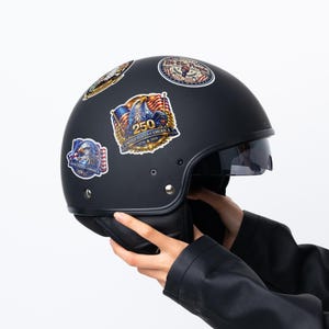K&ouml;nnte beinhalten: Ein mattschwarzer Motorradhelm mit Visier, verziert mit patriotischen Aufklebern. Die Aufkleber zeigen Adler, Flaggen und den Text "250 United States of America 1776-2026". Der Helm wird von einer Person gehalten, die eine schwarze Jacke tr&auml;gt.