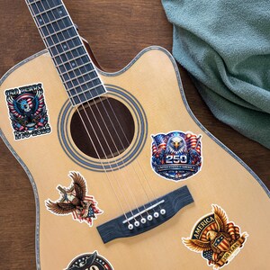 K&ouml;nnte beinhalten: Akustikgitarre mit patriotischen Aufklebern. Die Aufkleber zeigen Adler, amerikanische Flaggen und Text zur Feier von 250 Jahren der Vereinigten Staaten von Amerika, mit den Daten 1776-2026. Die Gitarre hat eine nat&uuml;rliche Holzoberfl&auml;che.