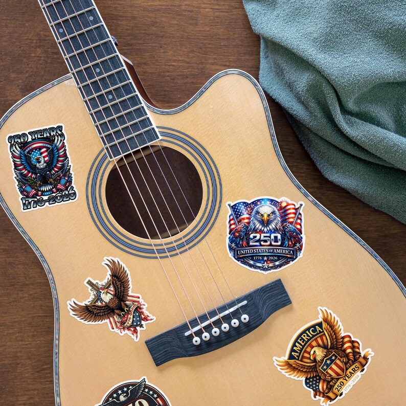 K&ouml;nnte beinhalten: Akustikgitarre mit patriotischen Aufklebern. Die Aufkleber zeigen Adler, amerikanische Flaggen und Text zur Feier von 250 Jahren der Vereinigten Staaten von Amerika, mit den Daten 1776-2026. Die Gitarre hat eine nat&uuml;rliche Holzoberfl&auml;che.