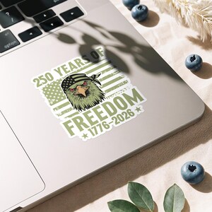 K&ouml;nnte beinhalten: Ein Aufkleber mit einem Adler, der ein Bandana mit dem Design der amerikanischen Flagge tr&auml;gt. Der Aufkleber hat den Text "250 YEARS OF FREEDOM 1776-2026" in Gr&uuml;n und Wei&szlig;, mit einem verwitterten Hintergrund der amerikanischen Flagge.