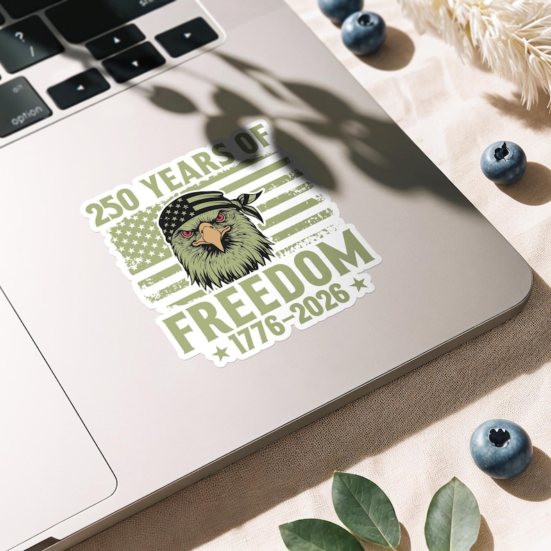K&ouml;nnte beinhalten: Ein Aufkleber mit einem Adler, der ein Bandana mit dem Design der amerikanischen Flagge tr&auml;gt. Der Aufkleber hat den Text "250 YEARS OF FREEDOM 1776-2026" in Gr&uuml;n und Wei&szlig;, mit einem verwitterten Hintergrund der amerikanischen Flagge.