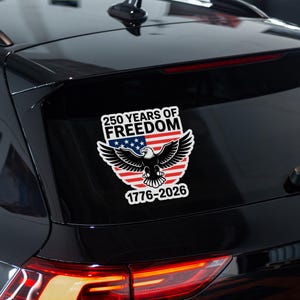 K&ouml;nnte beinhalten: Ein wei&szlig;er Aufkleber mit dem Text "250 YEARS OF FREEDOM" &uuml;ber einem Design der amerikanischen Flagge und einem Wei&szlig;kopfseeadler. Der Aufkleber enth&auml;lt auch die Daten "1776-2026". Der Aufkleber ist auf der Heckscheibe eines schwarzen Fahrzeugs angebracht.